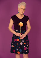 Woman holds flower in front of blue based knee length skirt with blue, orange, plum and pale pink splotch print, paired with a dark purple top