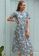 Woman wears midi length cotton dress with short sleeves, in coral, sage and lemon floral print on a mid blue base