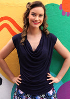 Woman standing front on with hands on hips wearing a cowl neck rayon navy blue cap sleeve top with floral bottoms.