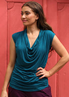 Woman standing in front of pink door wearing a cowl neck rayon teal cap sleeve top.