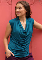 Woman wearing a cowl neck rayon teal cap sleeve top with maroon pants.