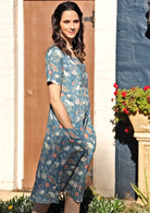 Woman stands side on in cotton over the knee dress in blue based floral print, with hands in pockets