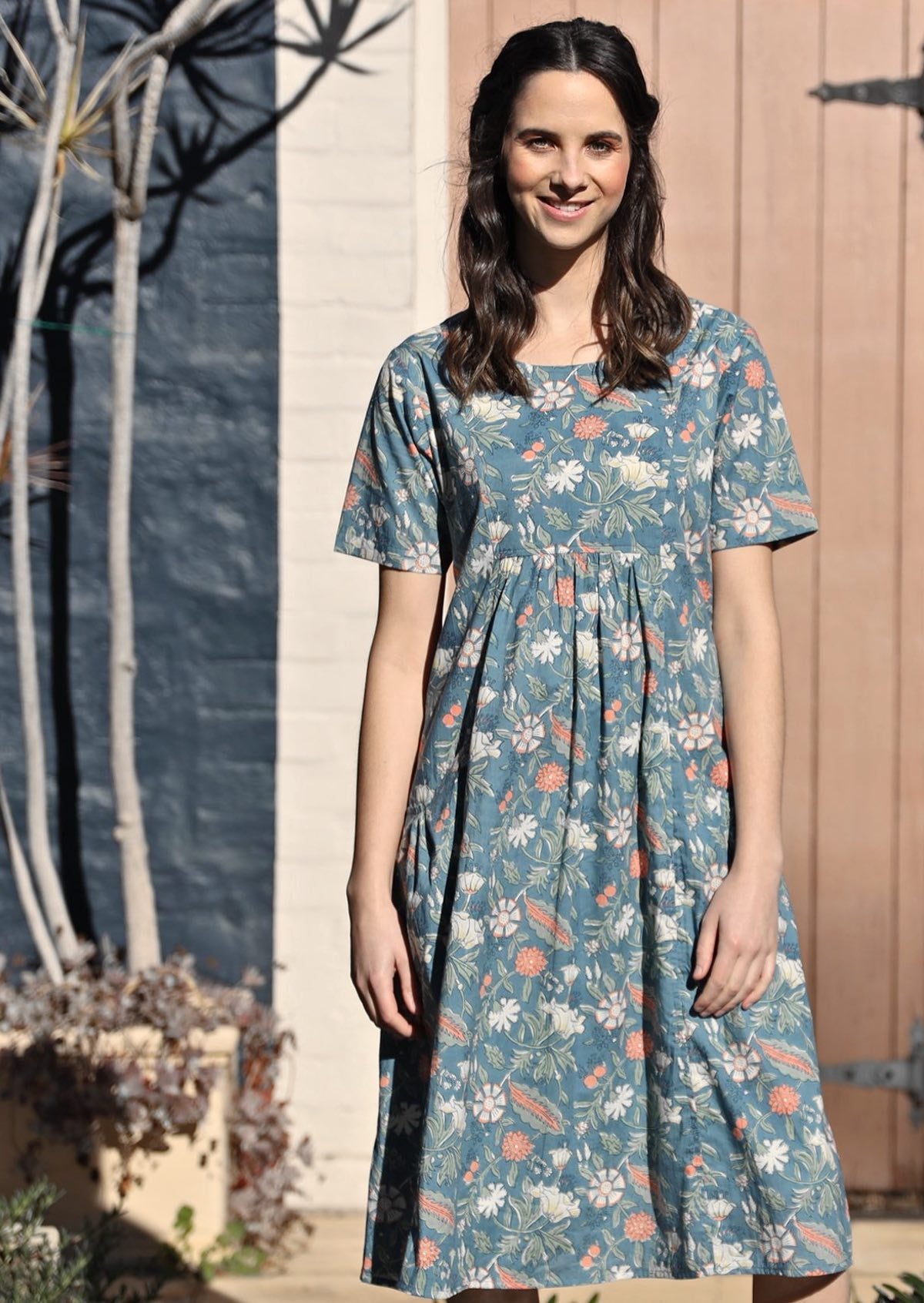 Woman wears over the knee length cotton dress with coral, sage and lemon florals on a mid blue base
