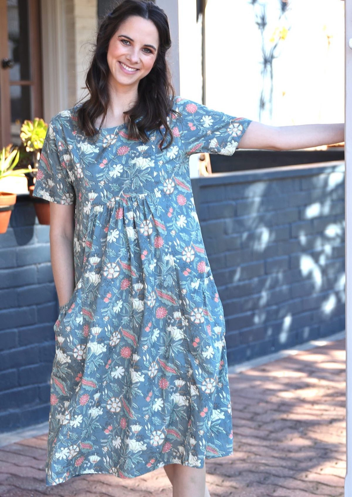 Woman leans from post, wearing floral print cotton sun dress that sits over the knee