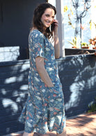 Woman walking away in dappled sunlight, wearing a cotton floral print dress with hand deep in pocket